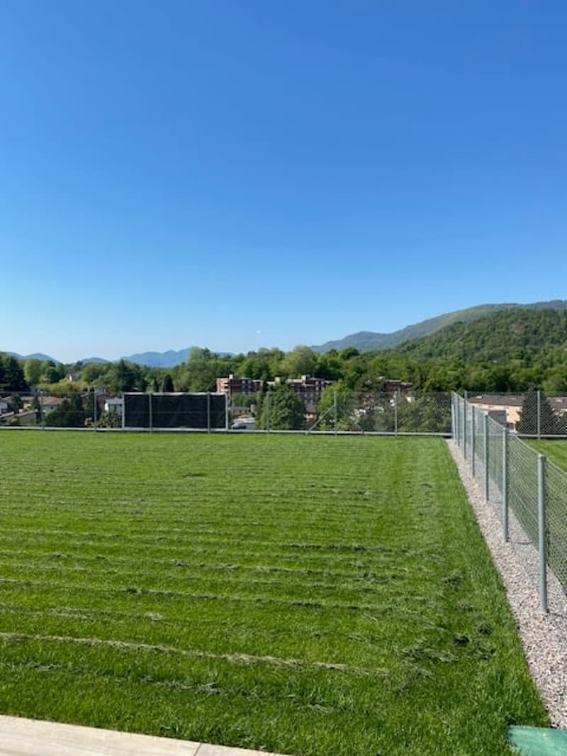 A large grassy field surrounded by a fence, with a paved area that appears to be a parking lot. The field is situated in a scenic area with mountains and trees visible in the background.