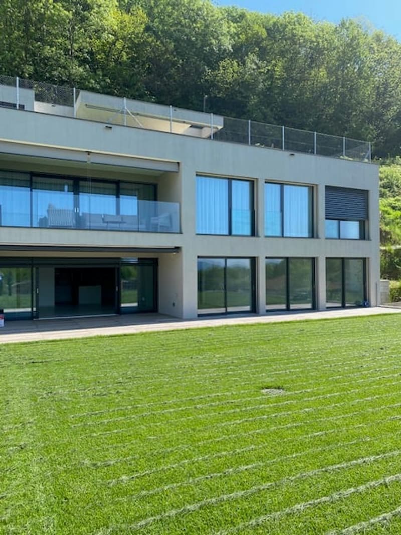 A modern, multi-story building with large glass windows and a balcony or terrace on the upper level. The building is surrounded by a well-manicured lawn.