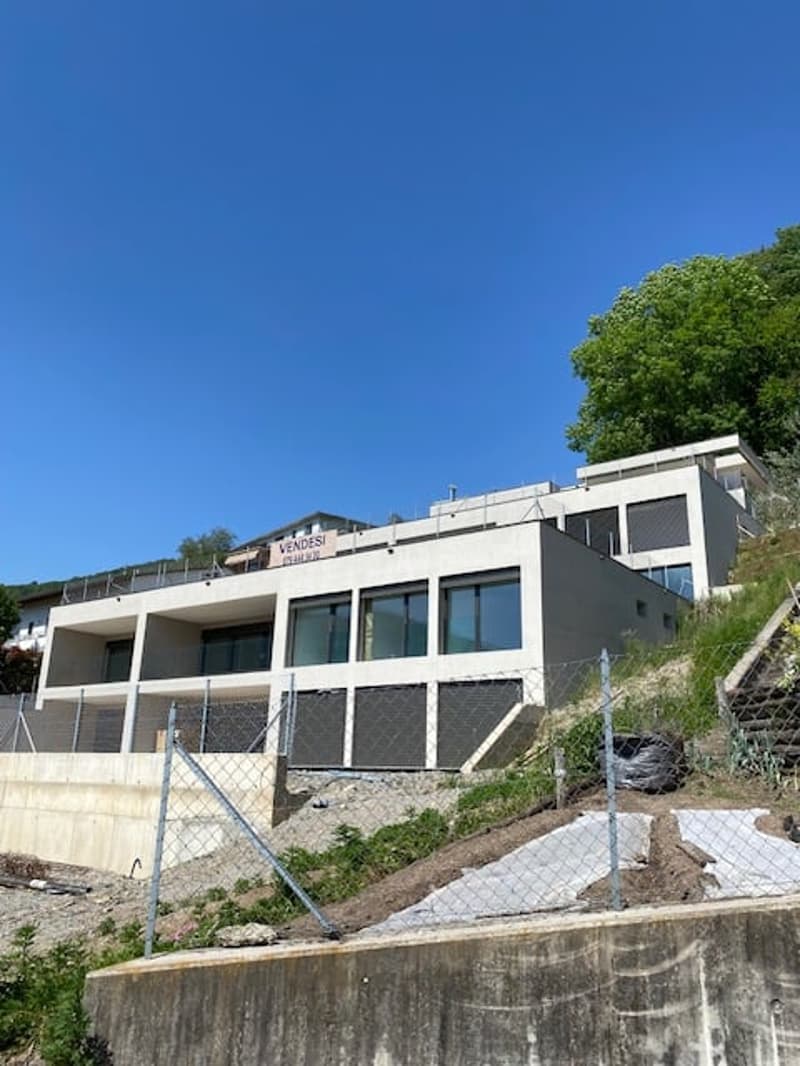 A modern, multi-story building with a balcony or terrace visible, situated on a hillside with lush greenery surrounding it. The building has a sleek, contemporary design with large windows and a gray exterior.
