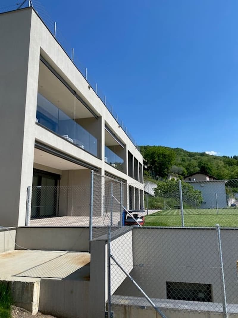 A modern multi-story building with large windows and balconies, surrounded by a fenced outdoor area with grass.