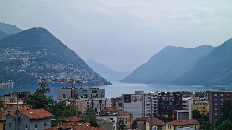 Aerial view, mountainous area, lake, city buildings, crane, hills, and a cloudy sky. Buildings in various colors, green trees and vegetation.