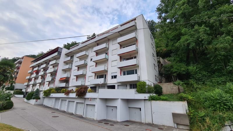 Multi-story apartment building, white exterior, multiple balconies, garages, greenery, street in front