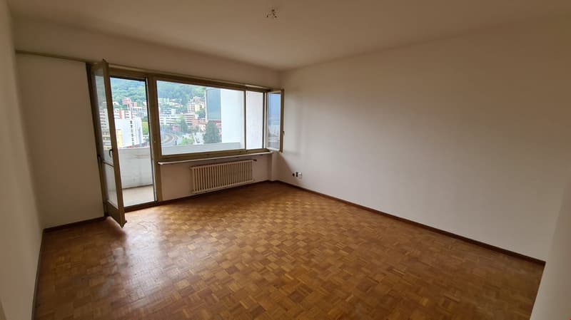 Large wooden floor, white walls, large windows leading to balcony, glass door, radiator, electrical outlet, empty room