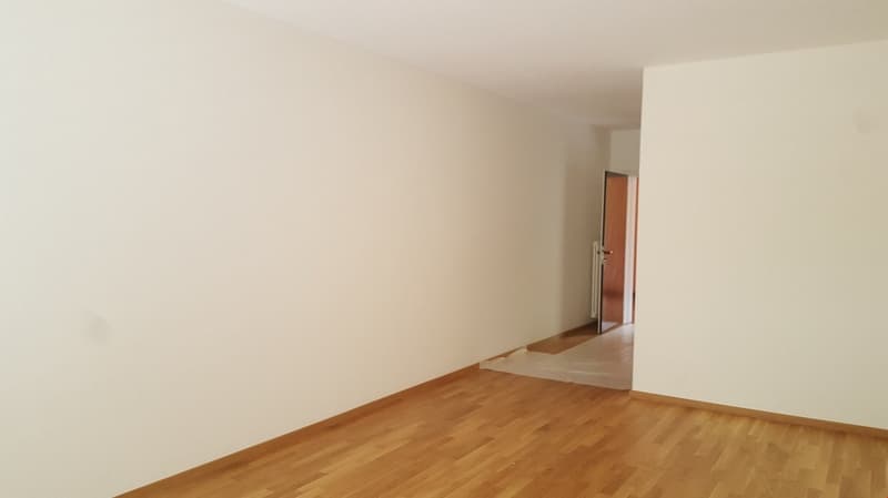 An empty room with white walls, wooden floors, and an open door leading to another room.