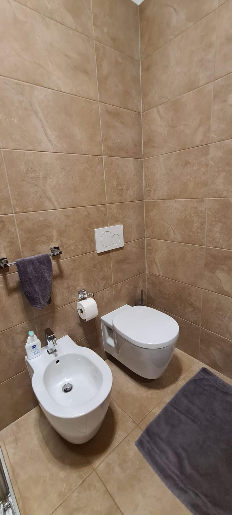 modern compact bathroom, beige tiled walls, bidet, toilet, floor-mounted sink, toilet paper holder, hand towel holder, small rug, cleaning products, bidet faucet