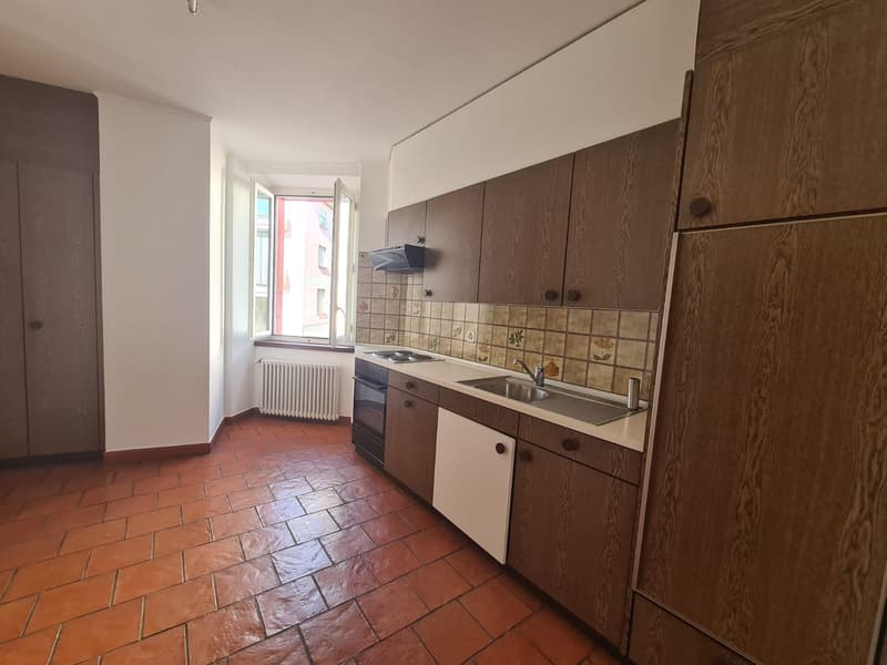 Brown wooden cabinets, tiled floor, stainless steel sink, gas stove, white tiled wall, radiator, window with a view of the outside