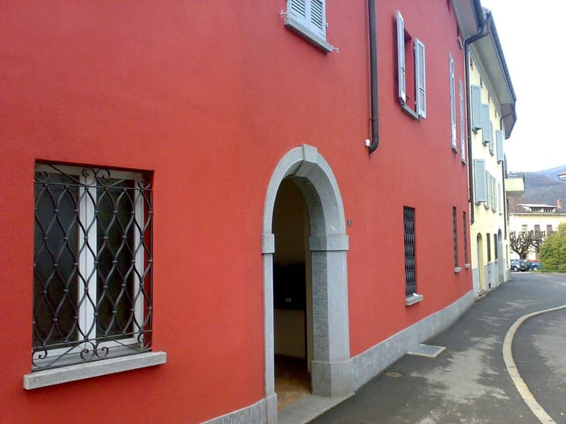A red building with a door at the bottom of an archway. The building has several windows with white shutters. A chimney is present. There is a narrow alley next to the building.