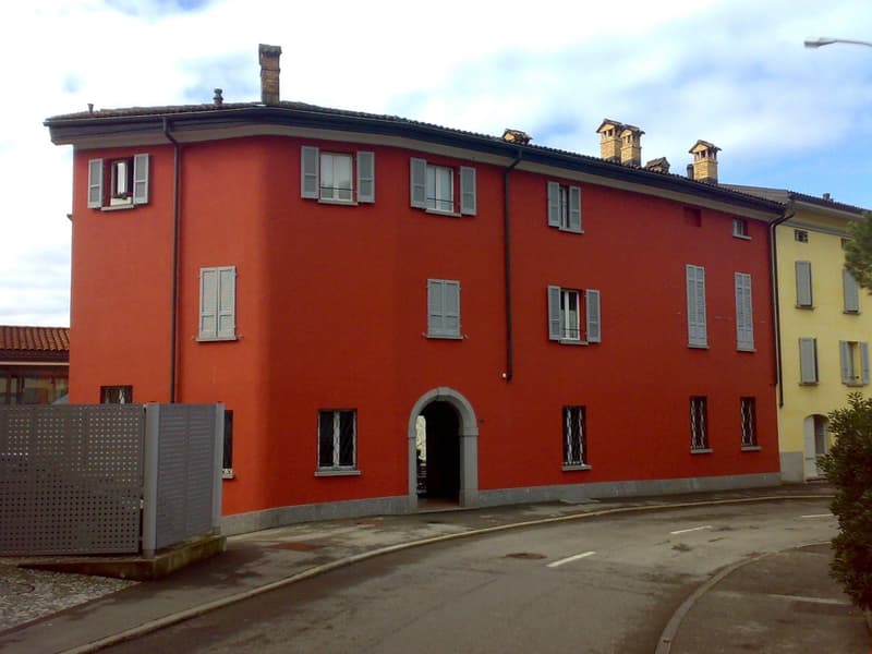 Multi-story building, red exterior, arched entryway, white shutters, multiple windows, chimneys, metal fence, yellow building nearby