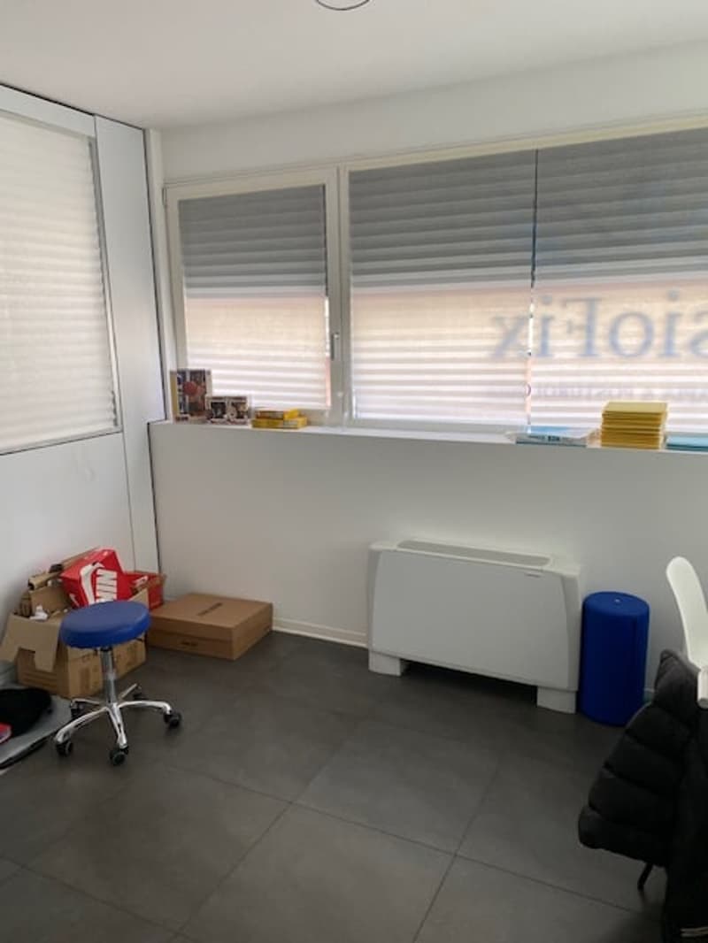Empty room with white walls, gray floor, two windows with blinds, a white radiator, a blue stool, a blue trash bin, a black chair, and some boxes on the floor.