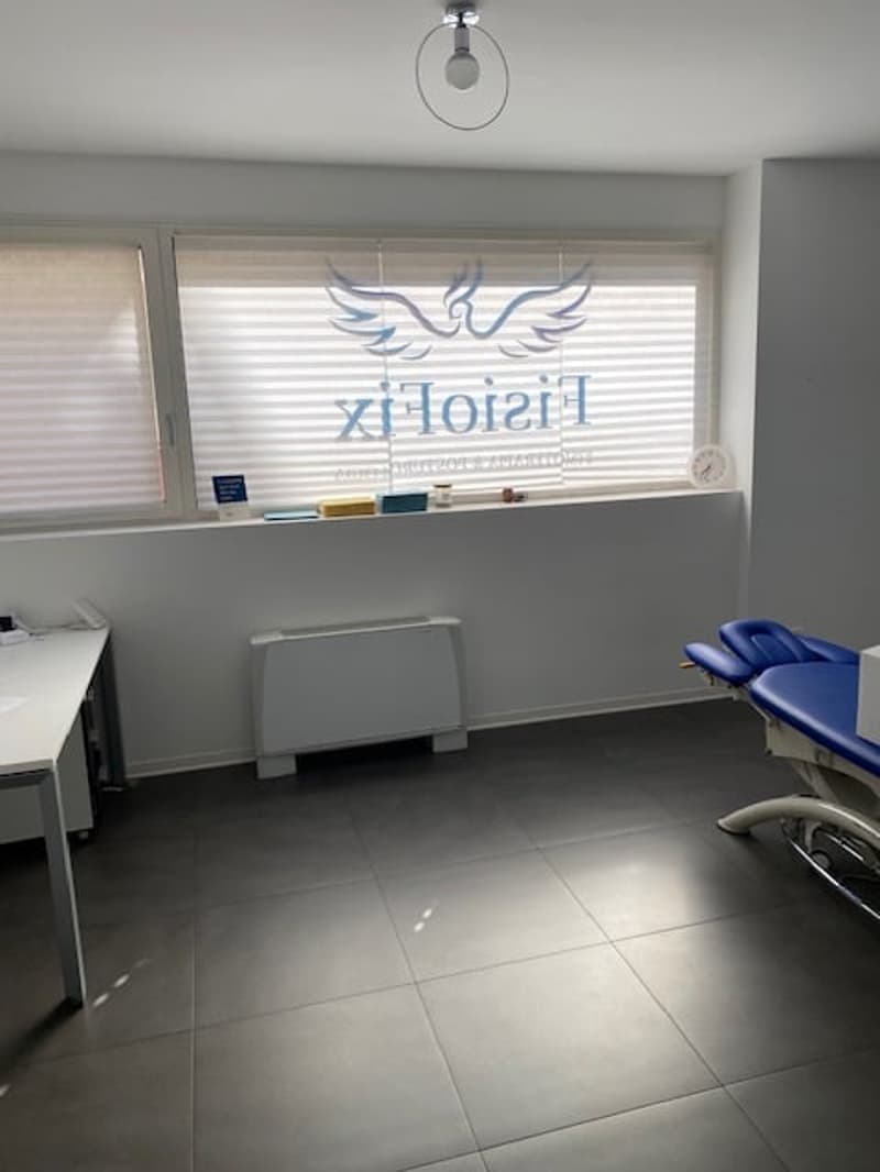 Empty room with white walls, gray floor, desk, blue chairs, window with blinds, heater, clock, and logo on the window