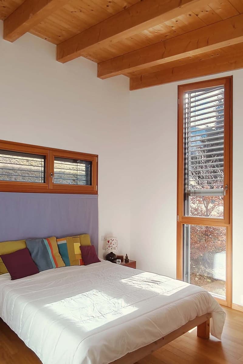 Bedroom with wooden bed, white bedding, wooden ceiling, wooden floor, large window, nightstand with lamp, decorative items