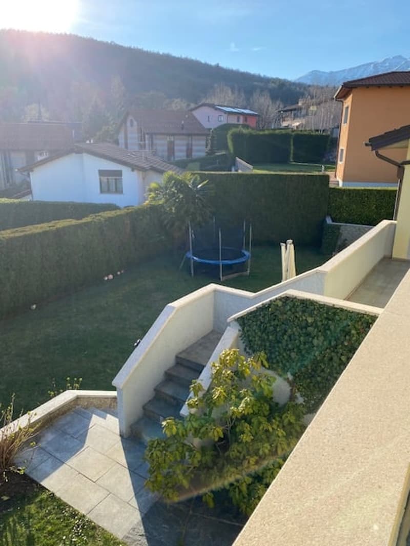 Aerial view of a garden with a trampoline, hedges, and a staircase leading to a balcony with a view of the mountains.