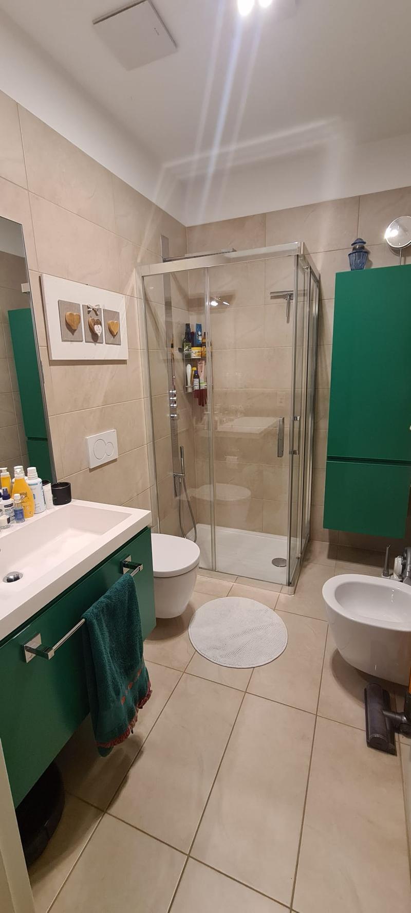 Modern bathroom with white tiles, glass shower enclosure, white vanity with sink, bidet, green cabinets, towel rack with towel, mirror on wall