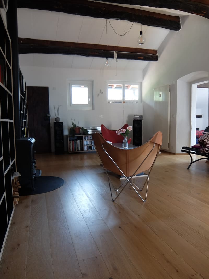 Living room with wooden floor, exposed wooden beams, two armchairs, a table, a bookshelf, a fireplace, and a window.