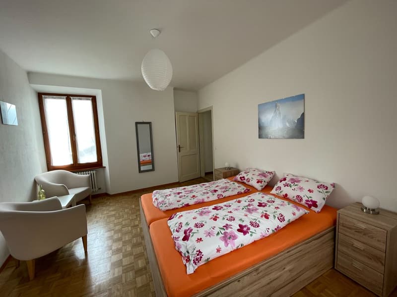 Bedroom with two beds, wooden floor, white walls, two nightstands, two chairs, radiator, mirror, lamp, picture, window