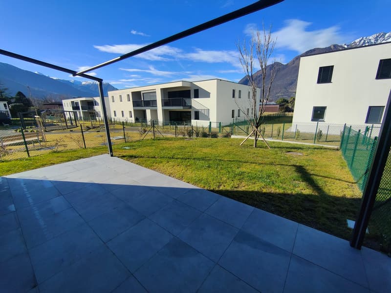 The image shows a modern apartment complex with several multi-story buildings surrounded by a well-maintained grassy area. The buildings have a minimalist, contemporary design with large windows and balconies. The complex appears to be situated in a sceni