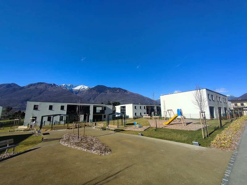 The image shows a residential complex with several multi-story buildings, a playground with swings and slides, and a grassy area with benches. The complex is surrounded by mountains in the background, creating a scenic setting.