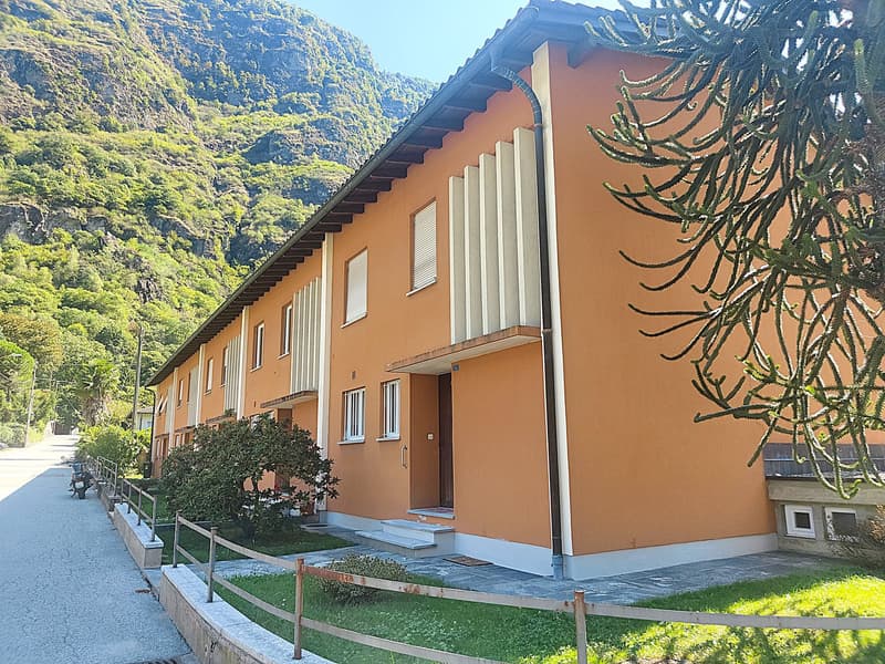 A multi-story residential building with an orange exterior, white trim, and a slanted roof. The building is surrounded by lush greenery and mountains in the background.