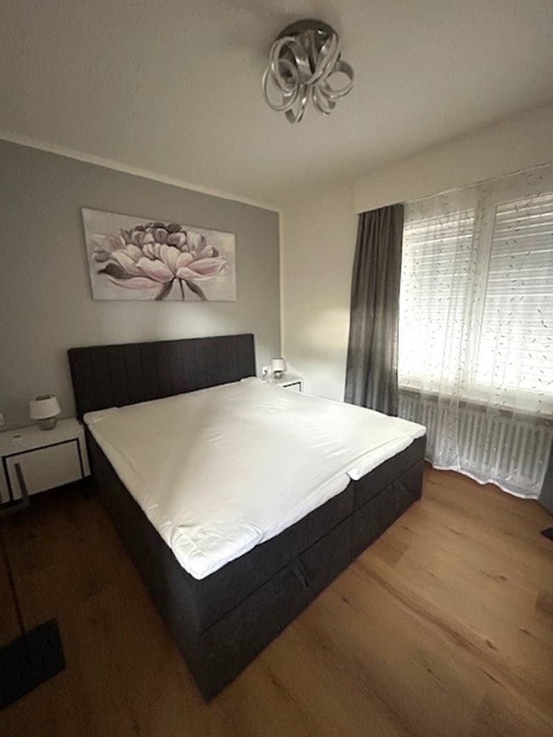 Bedroom with white sheets on bed, lamps on the side, white nightstands, white walls, ceiling lamp, painting on the wall, radiator under the window, wooden floor