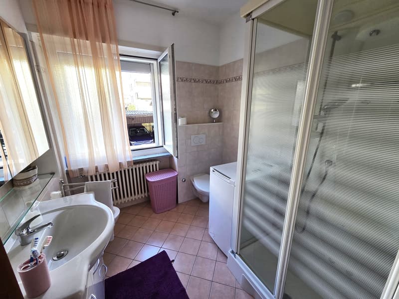 A bathroom with a sink, mirror, toilet, washing machine, shower, radiator, window, curtain, and a small basket on the mirror shelf