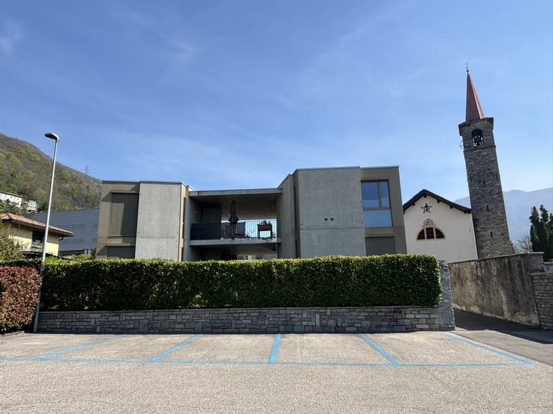Modern building with concrete exterior, large windows, balcony on the second floor, adjacent to a church with a tower, surrounded by a hedge and a parking lot