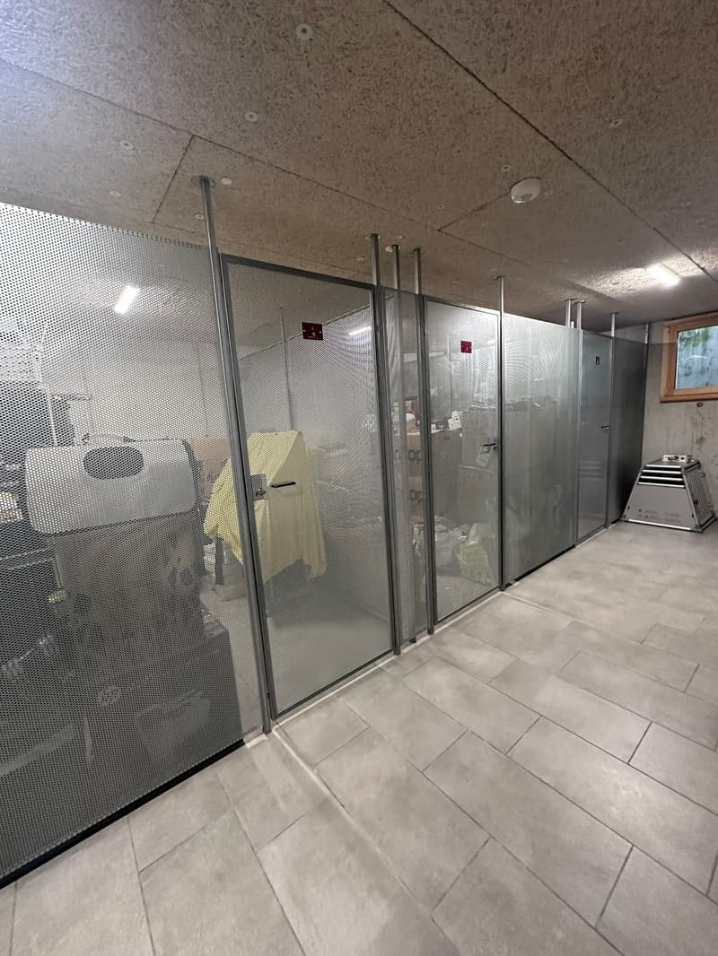 A room with tiled floor, metal mesh walls, and a glass door. Inside the room, there is a yellow cloth hanging on a metal pole, and a white object that seems to be a speaker. There are also some storage boxes and a white object that seems to be a heater on
