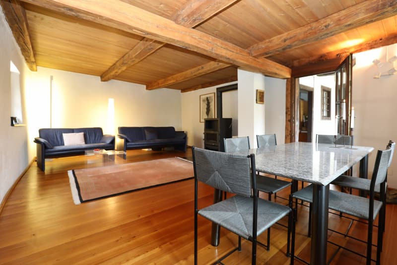 This image shows a spacious open-plan living and dining area with a high wooden ceiling and exposed beams. The living room features a pair of navy blue sofas, while the dining area has a large granite-topped table with black chairs. The room has a warm, r