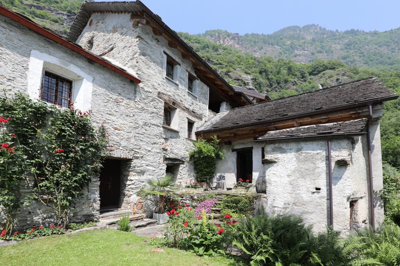 Old stone house with a tiled roof, surrounded by a lush garden with flowers and greenery. The house has a balcony or terrace, and there appears to be a fireplace visible as well.