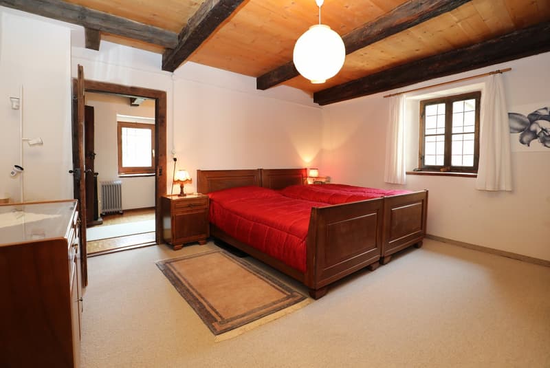 Spacious bedroom with wooden beams on the ceiling, a large wooden bed with a red comforter, wooden nightstands, and a window overlooking the room. The room has a cozy and rustic feel with the exposed wooden elements.
