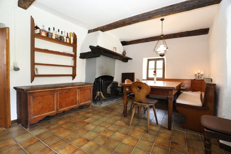 This image shows a rustic kitchen and dining area with exposed wooden beams, a fireplace, and a mix of antique and vintage furniture. The room has a tiled floor and features a wooden cabinet, a dining table with chairs, and various decorative items and ki