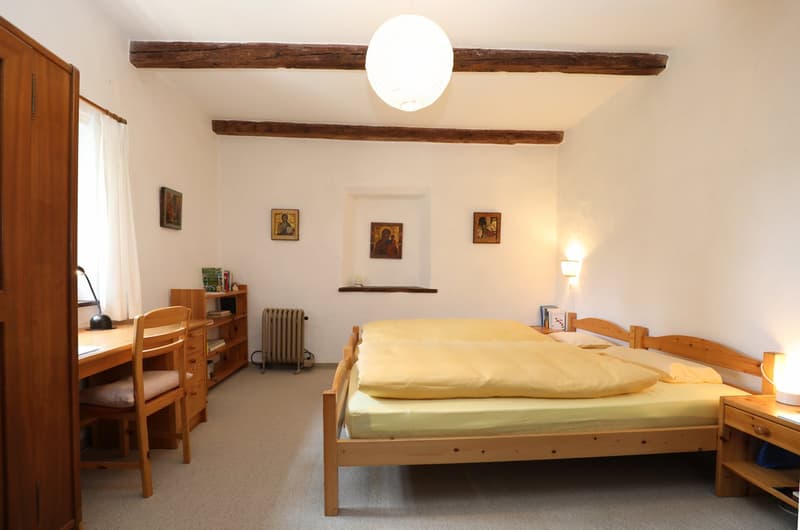 Cozy bedroom with wooden beams on the ceiling, wooden furniture including a bed, desk, and shelves, and religious artwork on the walls.