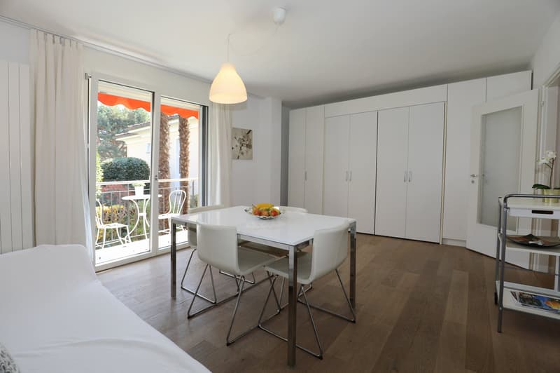 Dining area with a table and chairs, white walls, wooden floor, sliding glass door leading to a balcony with a view of the outside, a shelf with books and a flower vase, a white bed with a white cover