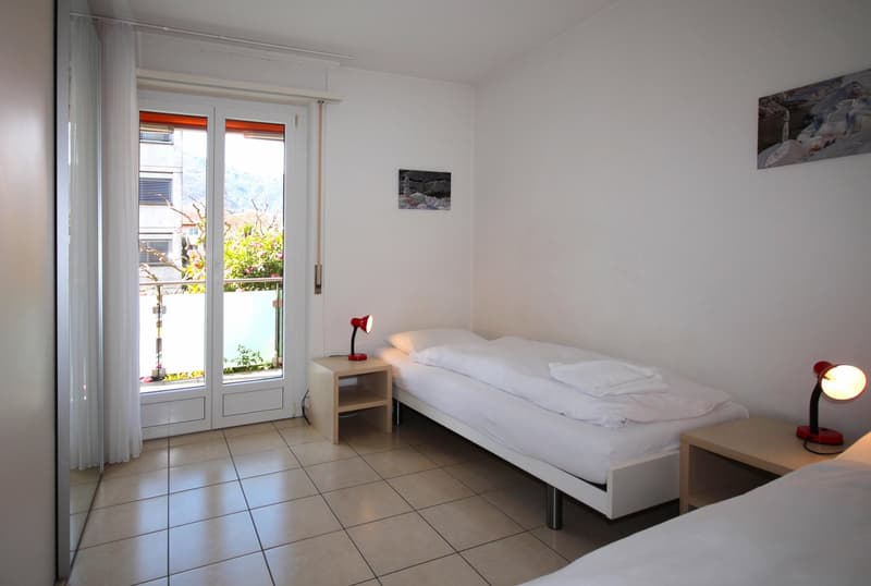 Two single beds, nightstands, lamps, white walls, tiled floor, sliding glass door, balcony, view of the outside
