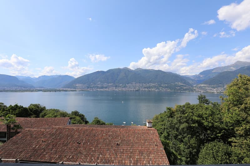 The image shows a scenic view of a lake surrounded by mountains and lush greenery. The lake appears to be Lake Como, a popular tourist destination in northern Italy. The image features a tiled roof in the foreground, suggesting a residential or commercial