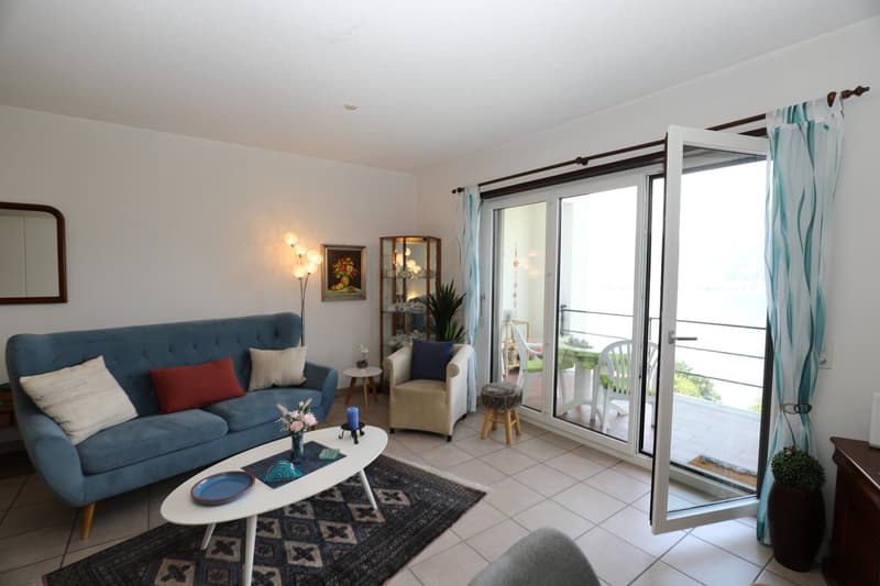 Spacious living room with a blue sofa, coffee table, and a balcony with a view. The room has white walls, tile flooring, and large windows that let in natural light.