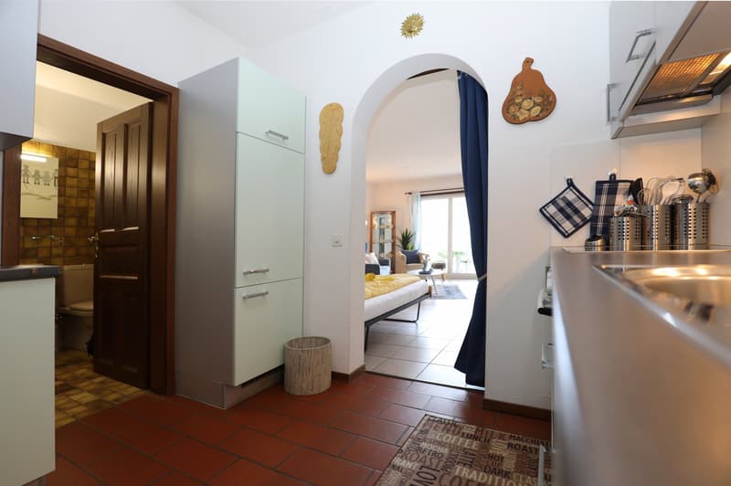 Spacious kitchen with wooden cabinets, stainless steel appliances, and a tiled floor. The kitchen opens up to a living room area through an arched doorway.
