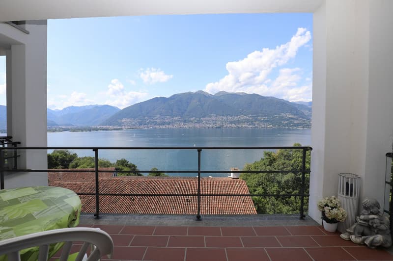 Spacious apartment with a large balcony overlooking a scenic lake surrounded by mountains. The balcony has a tiled floor and a wrought-iron railing. The view from the balcony is breathtaking, showcasing the tranquil lake, the distant mountains, and the cl