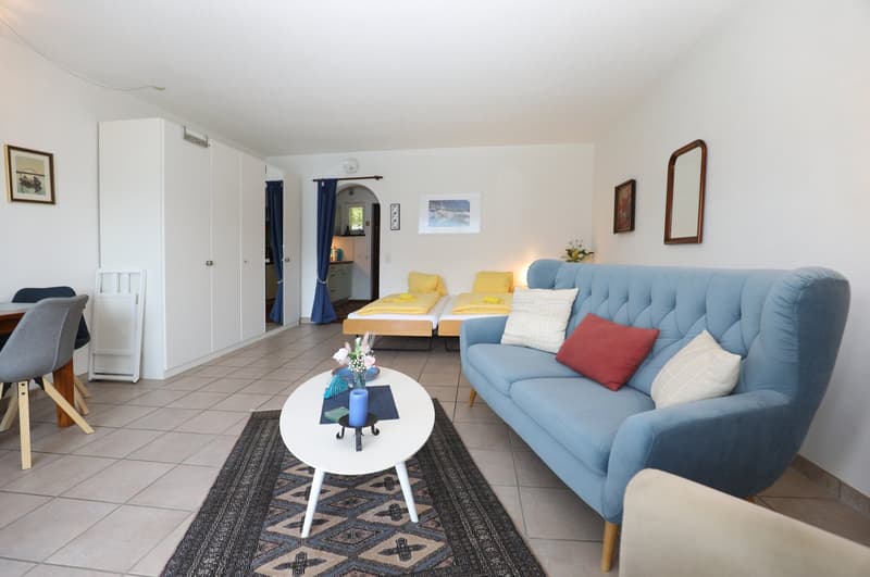 Spacious living room with tiled flooring, white walls, large blue sofa, coffee table, and a small balcony visible through the doorway.