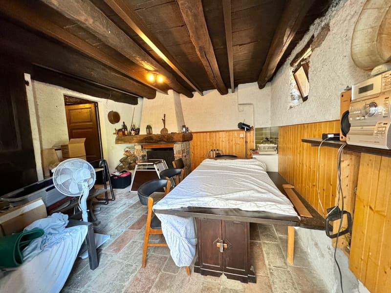 The image shows a rustic studio apartment with exposed wooden beams on the ceiling, a stone fireplace, and a kitchenette area with appliances. The room has a bed, chairs, and other furnishings, giving it a cozy and lived-in feel.