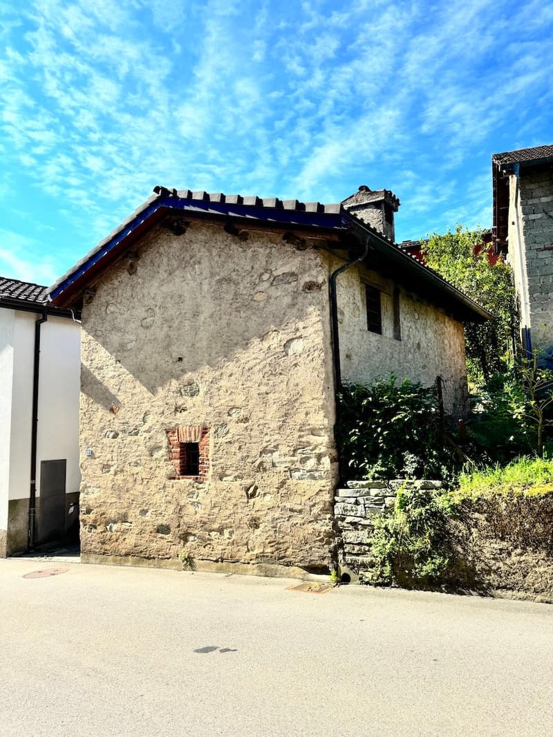 Old stone building with a tiled roof, surrounded by greenery and a paved area in the foreground.