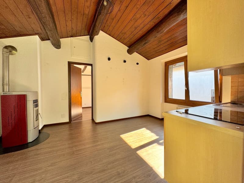 Open-concept studio apartment with wooden ceiling beams, hardwood flooring, white walls, and a yellow kitchen counter. The room features a fireplace and large windows providing natural light.