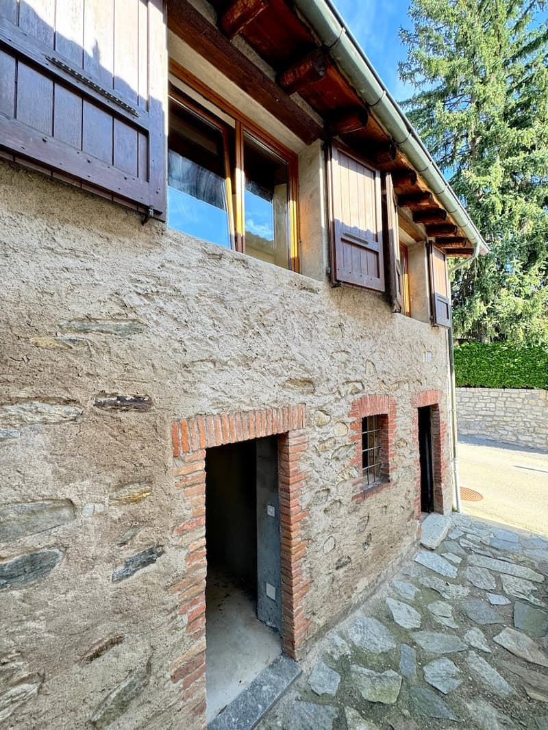 Two-story wooden house with stone walls, wooden shutters, and a covered porch. The house has a rustic, traditional design with a sloped roof and exposed wooden beams.