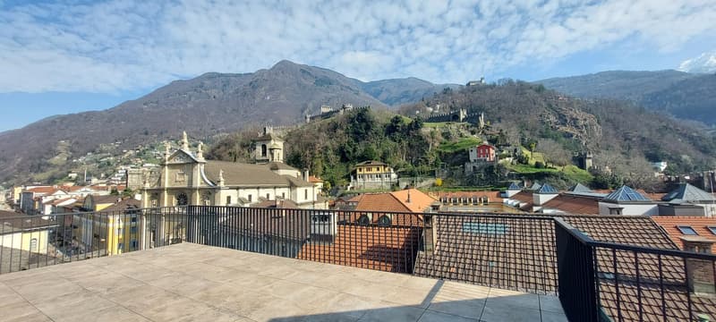View from an elevated terrace, overlooking town with tiled roofs and mountains in the background.