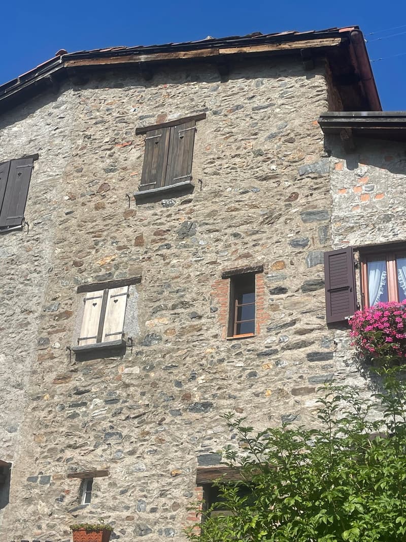 The image shows an old stone building with wooden shutters and windows. The building has a slanted roof and appears to be in a rural or mountainous area, with some vegetation visible in the foreground.