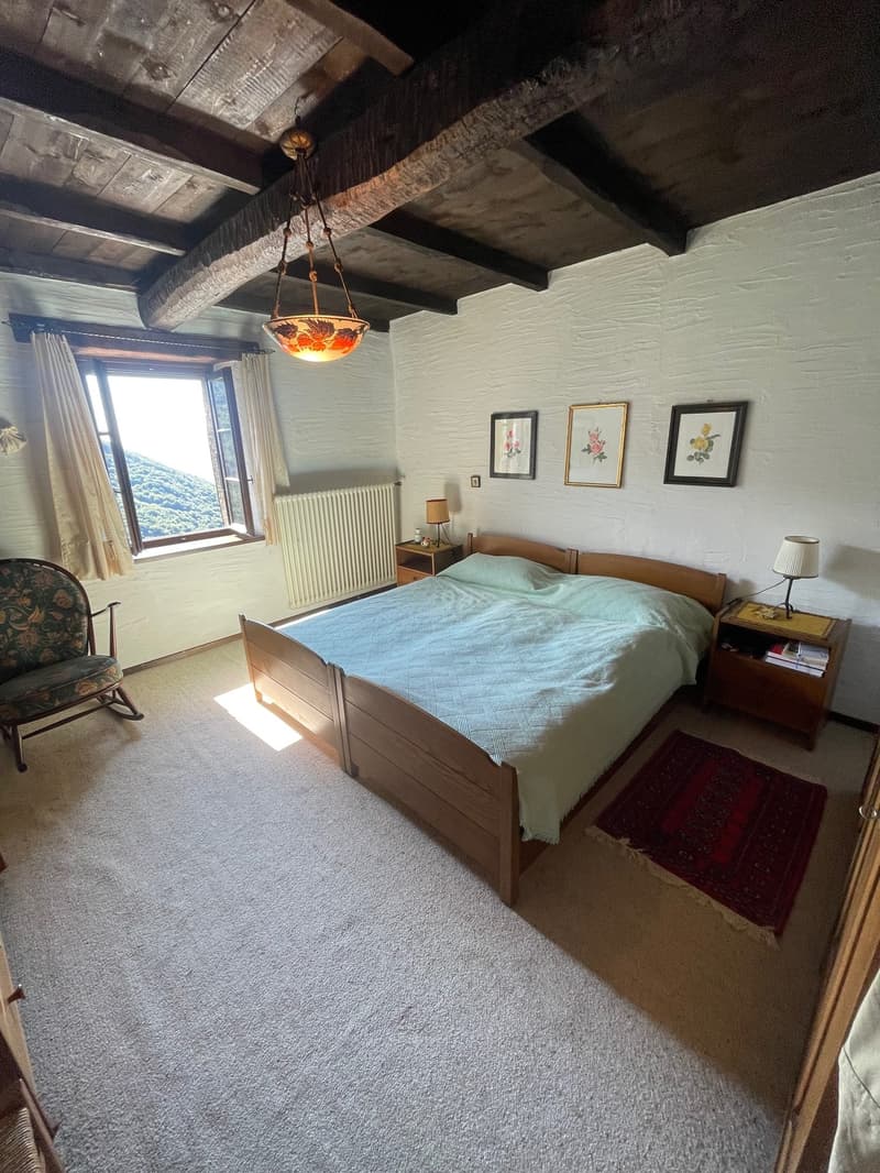 Spacious bedroom with wooden beams on the ceiling, a large window overlooking the landscape, a wooden bed frame, nightstands, and framed artwork on the walls.