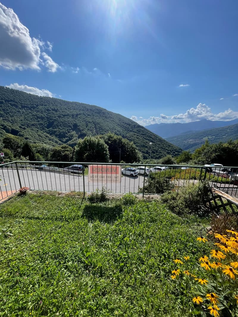 The image shows a scenic mountain landscape with lush green vegetation, including trees and a grassy area with yellow flowers. There is a parking area with a metal railing in the foreground, and the background features a blue sky with some clouds. The ove