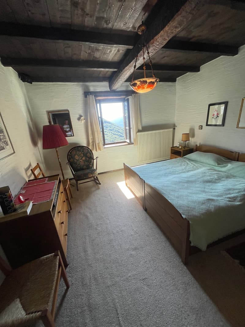 Rustic bedroom with exposed wooden beams on the ceiling, a large window with a view, a rocking chair, and a wooden dresser. The room has a cozy and warm atmosphere.