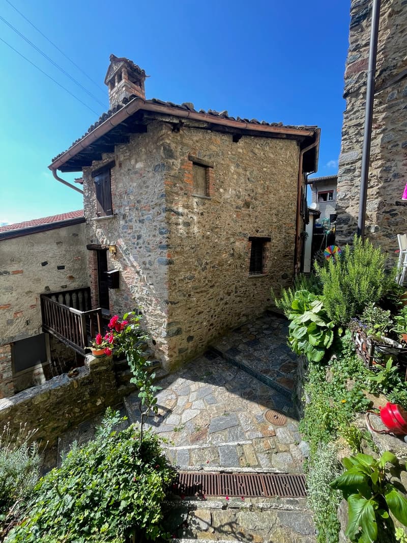 Two-story stone house with a tiled roof, balcony, and a lush garden with flowers and plants surrounding the entrance.