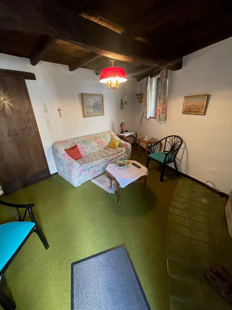 Cozy living room with a floral patterned sofa, a red lamp shade, wooden beams on the ceiling, and a fireplace. The room also has a small table with a chair, and some artwork on the walls.