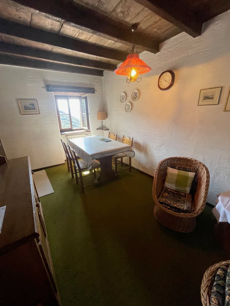 white brick walls, wooden ceiling, dining table with 6 chairs, wicker chair, radiator, wooden cabinet, framed pictures on wall, lamp, window, green carpet, clock